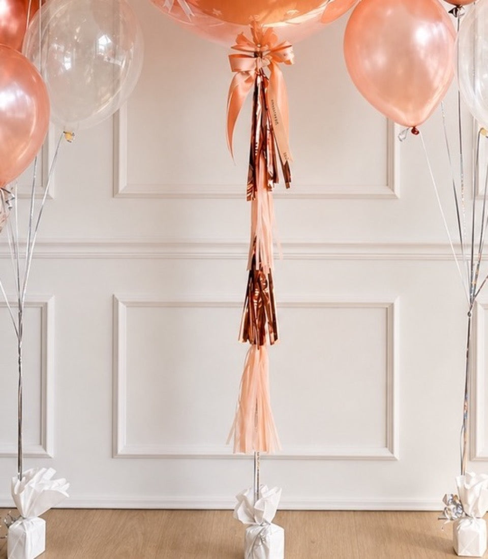 Rose gold balloon with tassel tail and ribbon bow, surrounded by matching pearl balloons against white paneled wall.
