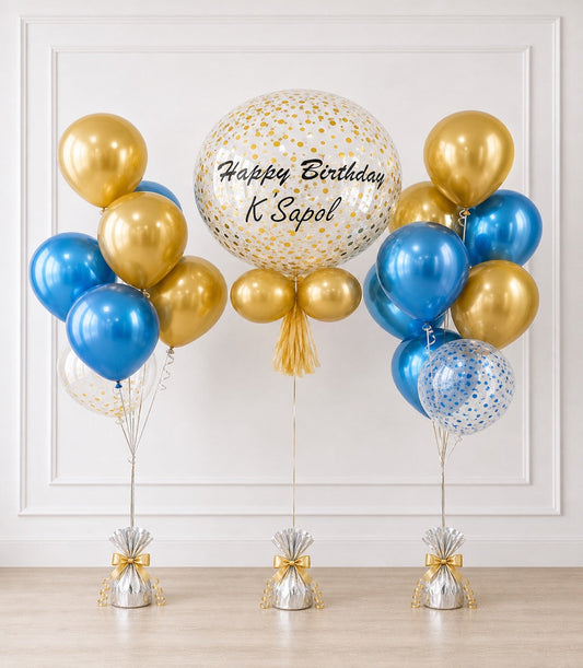 Gold and blue balloon bouquet with "Happy Birthday K Sapol" text on large confetti balloon, tied with gold tassels and weighted bases.