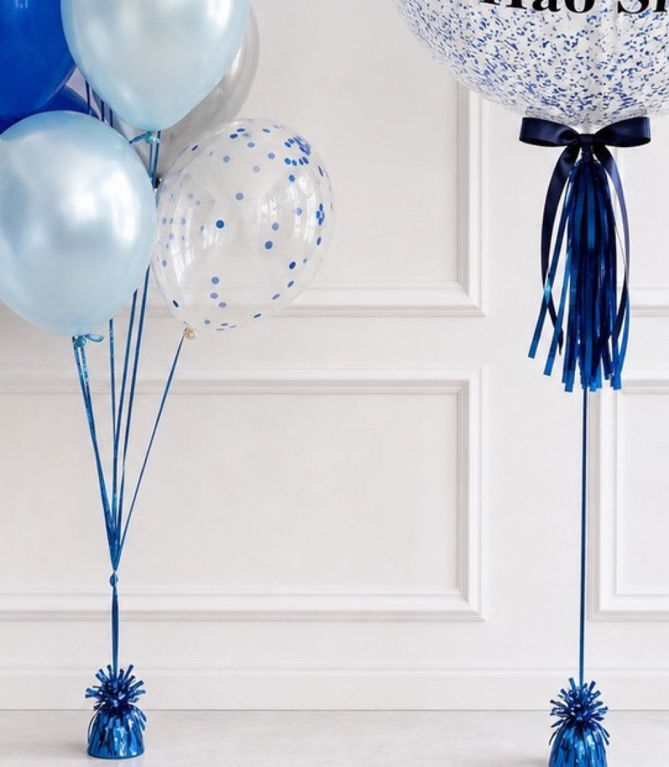 Blue and white balloon bouquet set with confetti-filled spheres, metallic weights, and ribbon ties against white paneled wall.