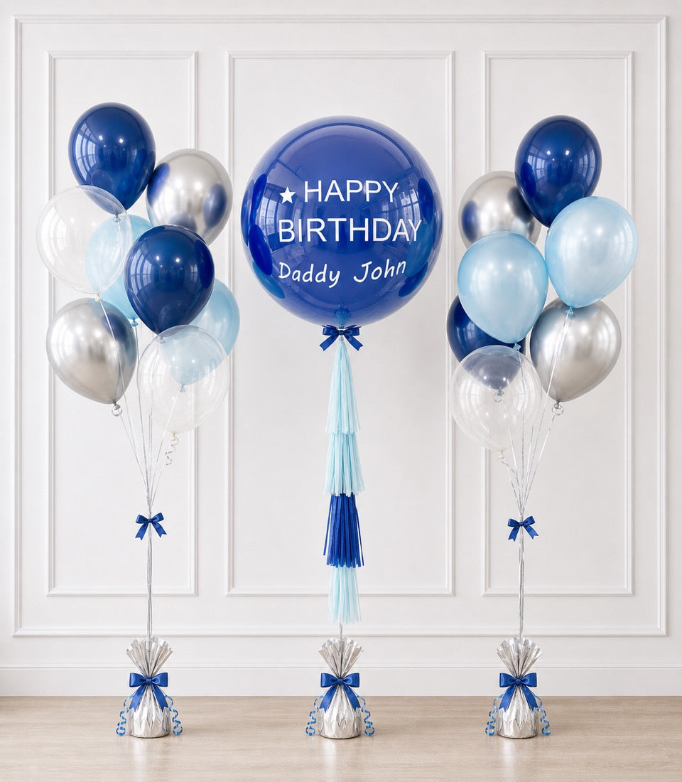 Blue and silver birthday balloon bouquet with personalized large centerpiece balloon reading "Happy Birthday Daddy John" against white paneled wall.