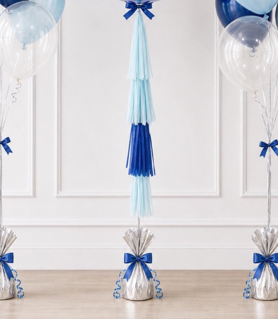 Blue and white balloon bouquets with matching tassels and ribbon bows displayed against a white paneled wall.