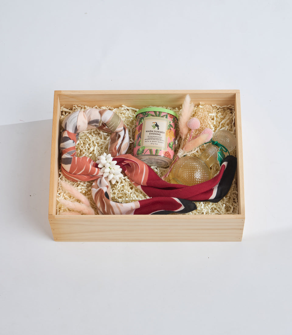 Wooden gift box containing a scented candle, decorative ribbons in coral and burgundy, bath items, dried flowers, and packing straw on white background.