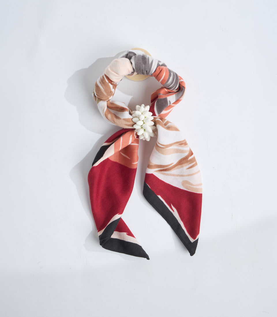 Colorful fabric scarf with geometric patterns in red, black, beige, and white, adorned with a small white flower charm on a wooden ring.