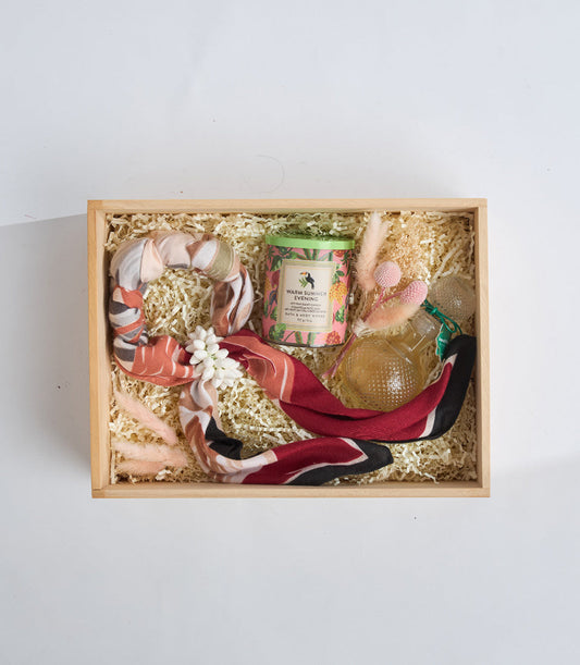 # Alt Text

Wooden gift box with natural straw fill containing a scented candle, patterned fabrics in coral and burgundy tones, and decorative items in gold and pink.