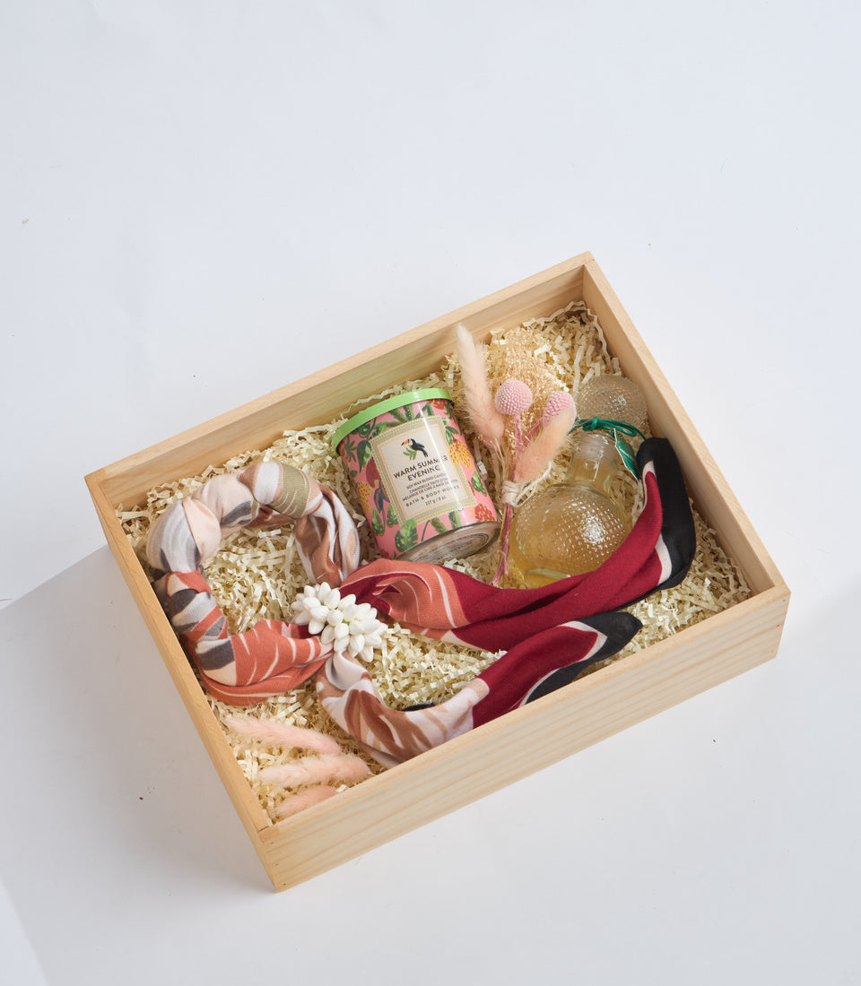 Wooden gift box filled with straw containing pink fabric flowers, decorative candle, glass perfume bottle, burgundy fabric, and beige crocheted items on white background.