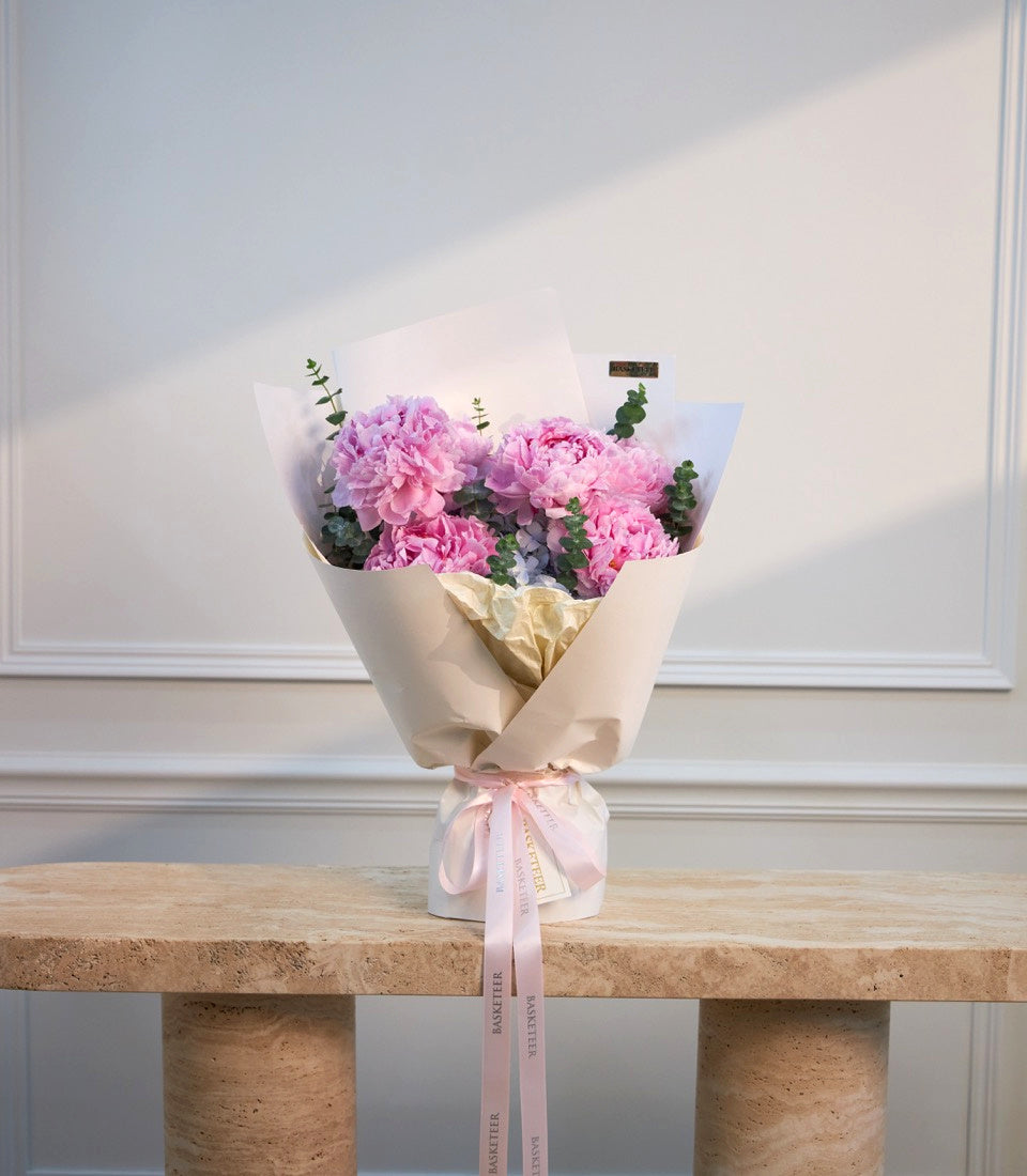 Pink peonies in cream and gold wrapped bouquet with green foliage and pink ribbon, displayed on natural stone bench against white wall.