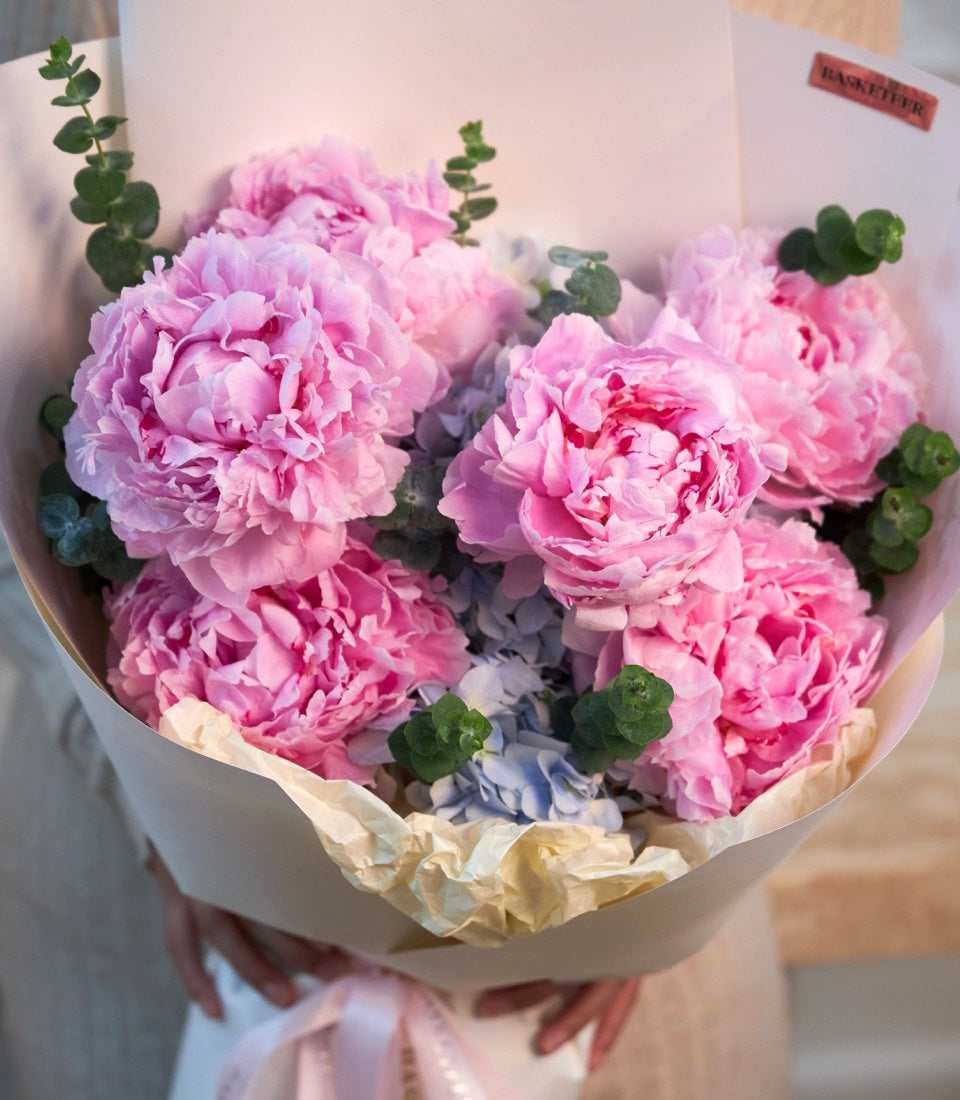 Luxurious bouquet of pink peonies with green eucalyptus stems, wrapped in white paper with cream tissue and pink ribbon.