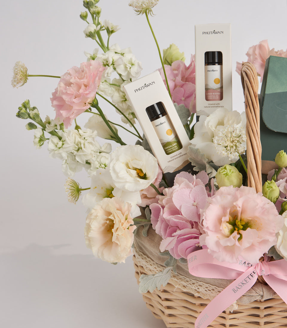 Phutawan spa gift set with aromatherapy bottles displayed in wicker basket surrounded by pink and white carnations, cream ribbon, and green leather pouch.