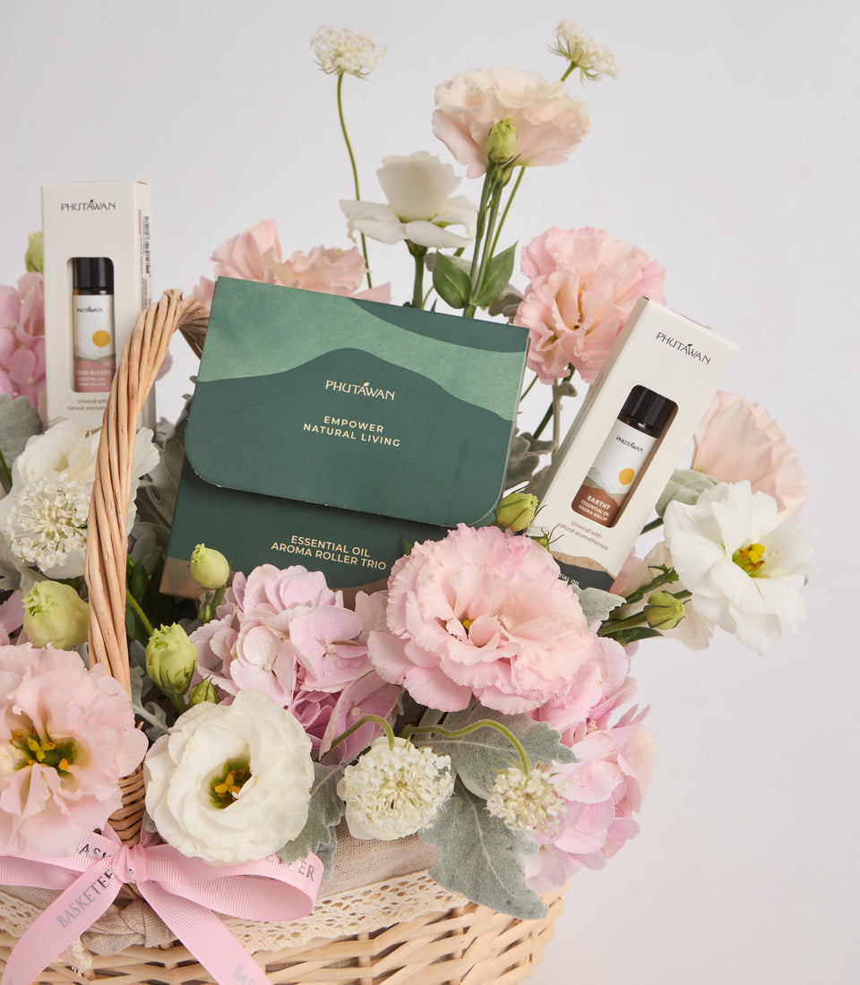 Phutawan essential oil aroma roller trio gift set with green packaging, displayed in wicker basket surrounded by pink and white carnations and eucalyptus.