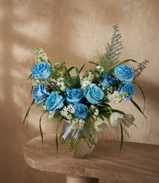Blue roses and white filler flowers in ribbed glass vase with white ribbon