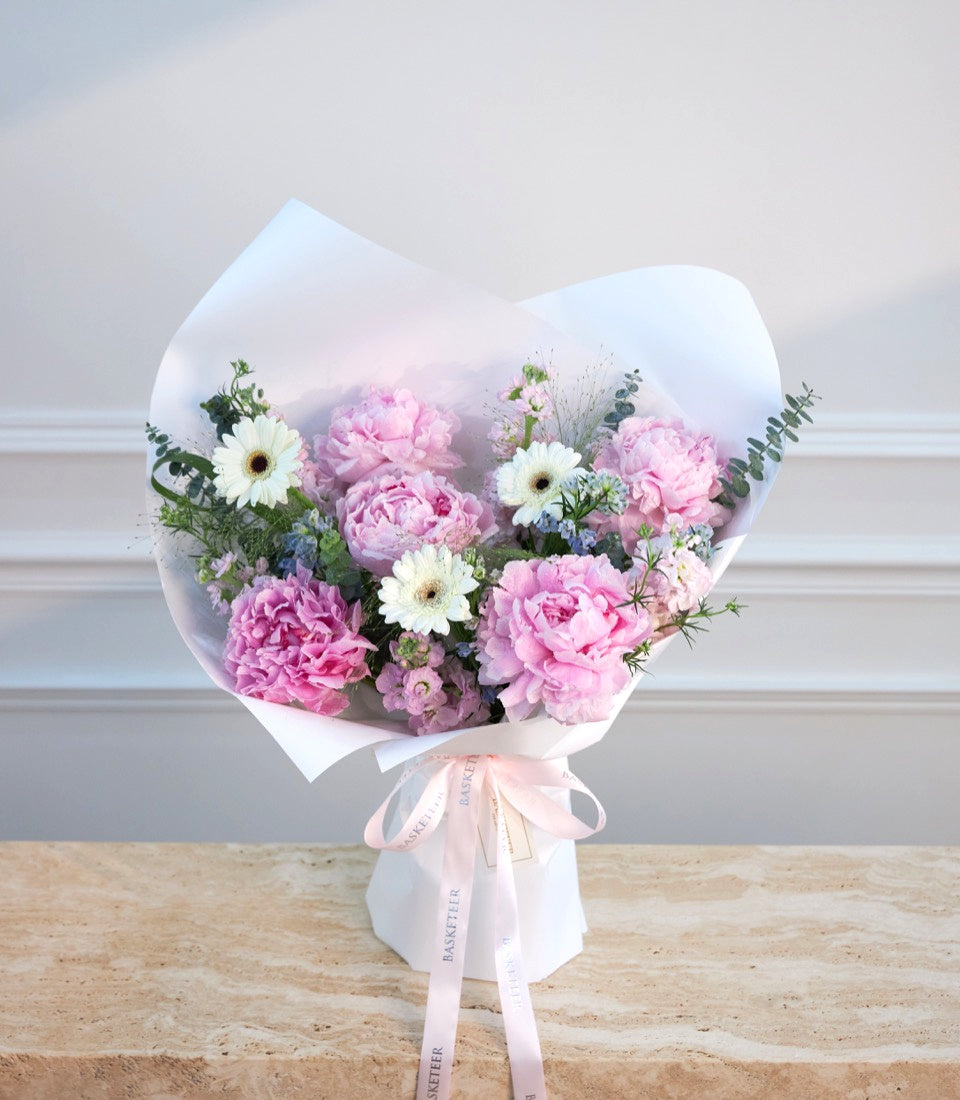 Elegant bouquet with pink peonies, white gerberas, and purple flowers wrapped in white paper with pink ribbon, displayed against a neutral background.