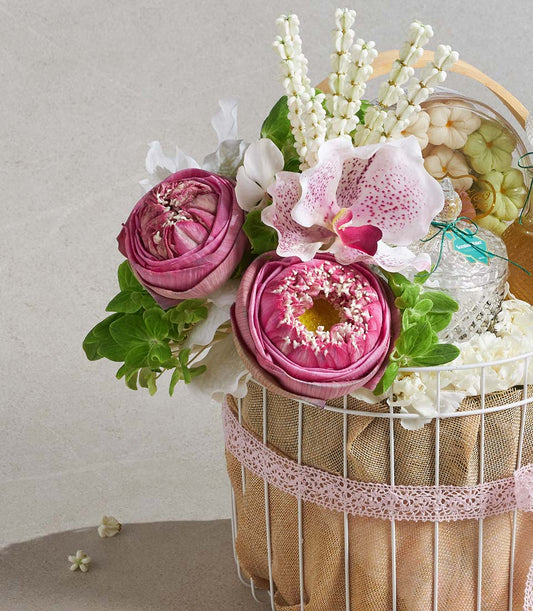 Pink lotus and orchid arrangement in white wire basket with cream confections and artisan treats