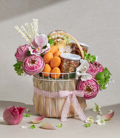 Elegant white wire gift basket with pink lotus, orchids, fresh fruits, and gourmet snacks tied with pink lace ribbon