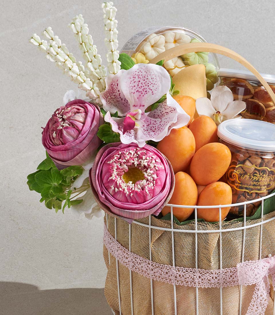 White wire gift basket with pink lotus flowers, fresh fruit, and gourmet treats in elegant pink and white arrangement