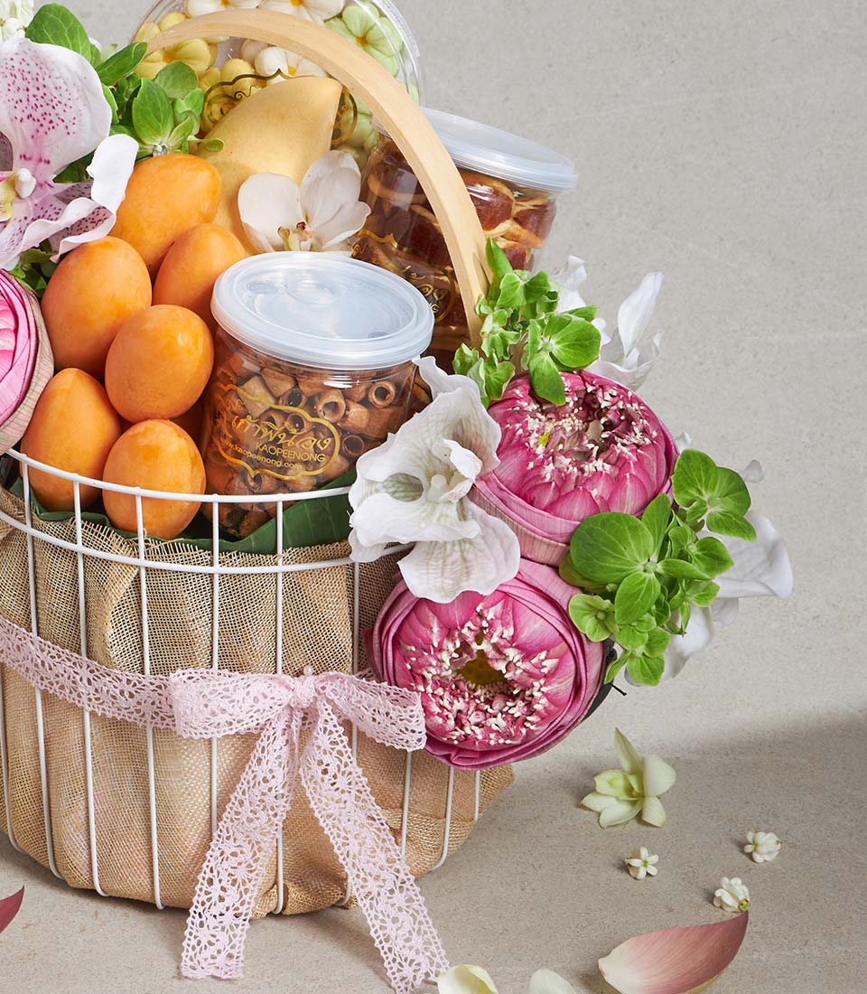 Luxe white wire gift basket with pink lotus flowers, orchids, fresh fruits, and gourmet snacks tied with pink lace ribbon