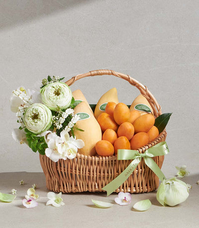 Pale yellow mangoes and orange stone fruit in woven wicker basket with white lotus and orchid bouquet, light green ribbon bow
