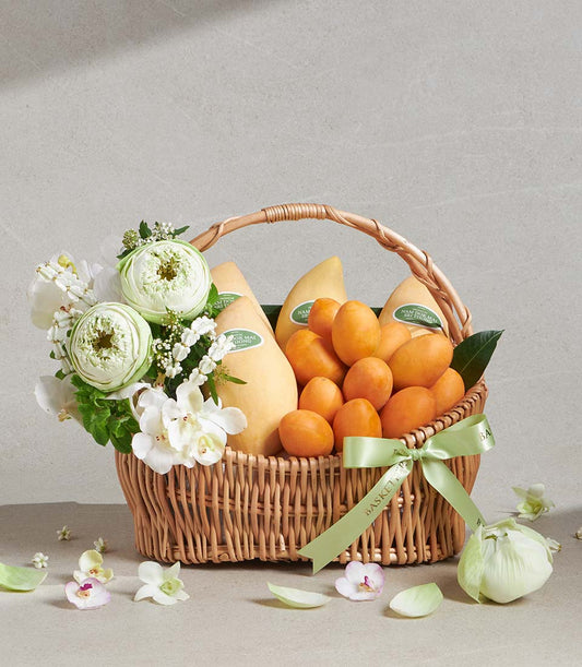 Pale yellow mangoes and orange stone fruit in woven wicker basket with white lotus and orchid bouquet, light green ribbon bow