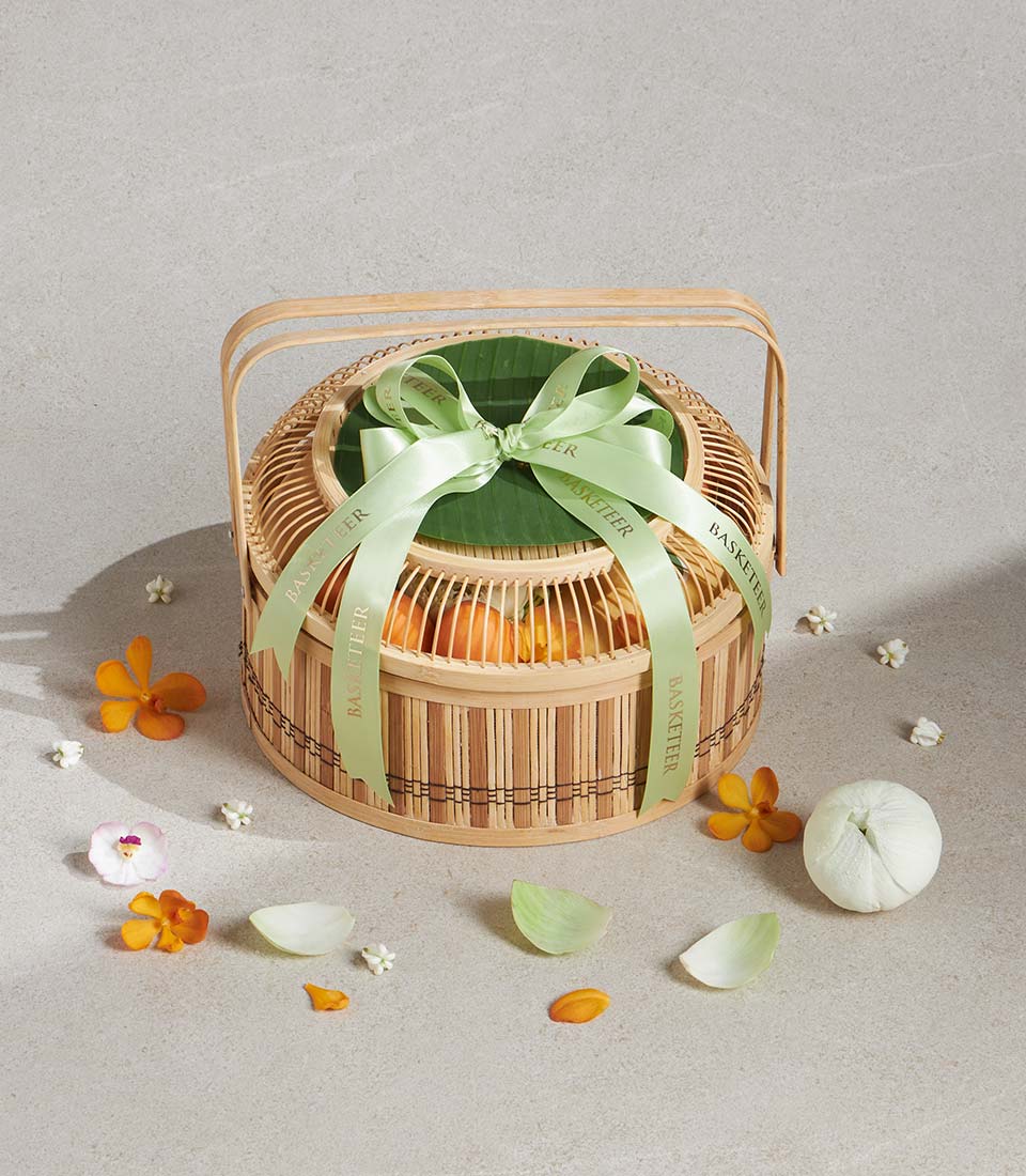 Two-tiered bamboo gift basket with citrus fruits, green ribbon bow, and jasmine and orchid florals for Songkran celebration