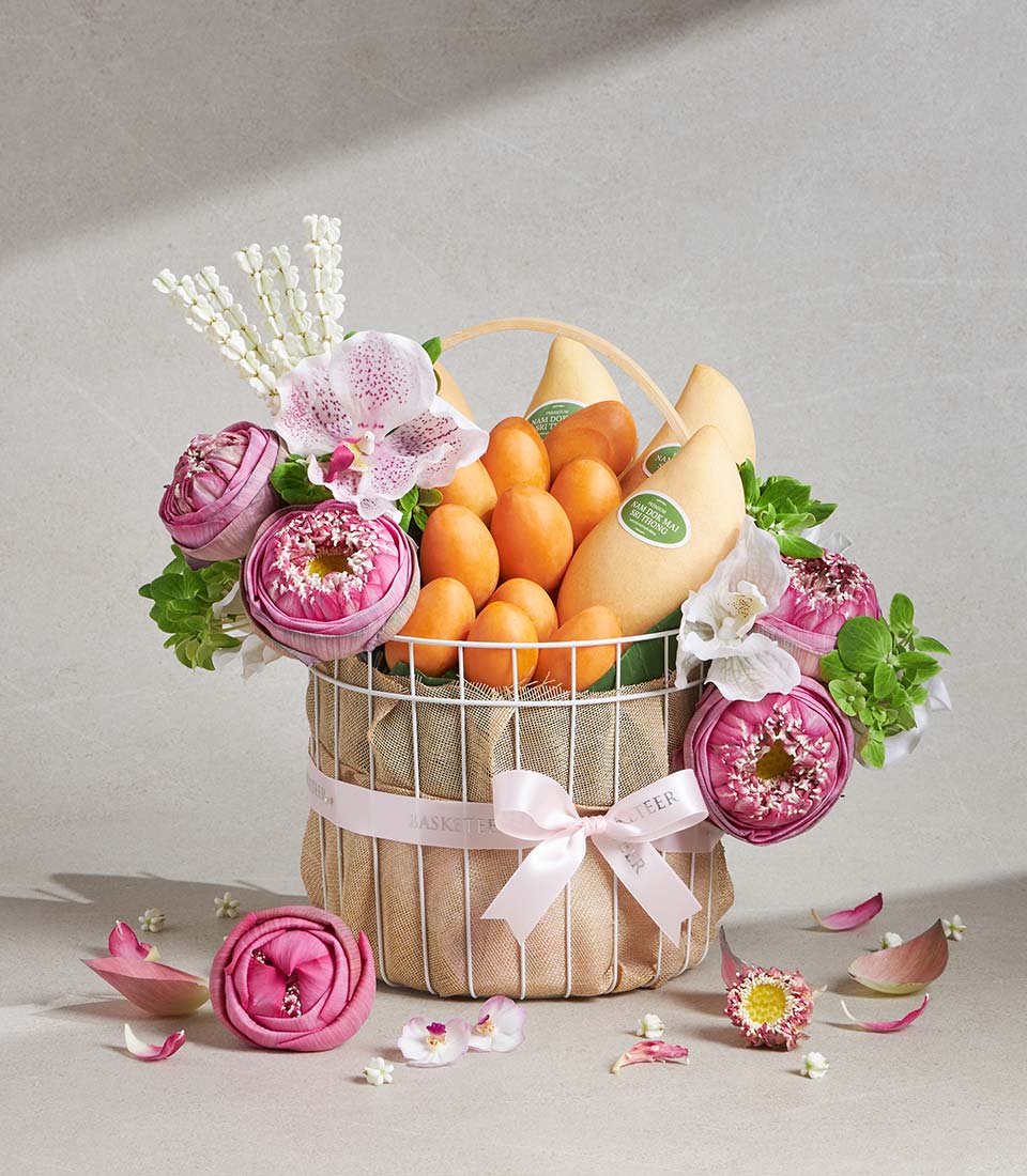 Premium white wire gift basket with fresh pink lotus, orchids, yellow mangoes and tropical fruits, tied with pink satin bow