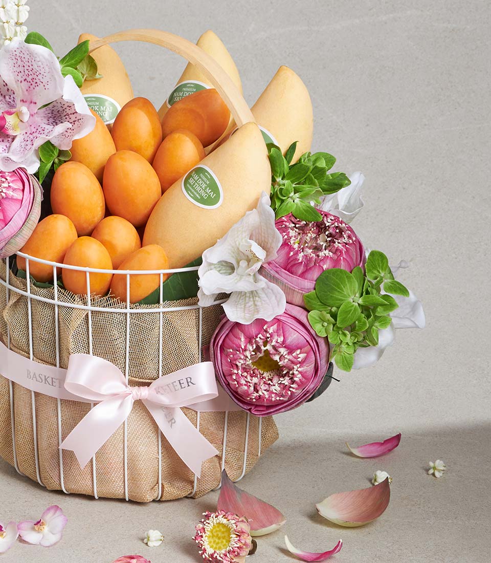 Pale yellow mangoes and orange fruits in white wire basket with fresh pink lotus, white orchids, and green foliage tied with
