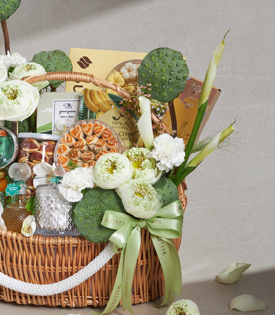 Woven wicker gift basket with lotus flowers, gourmet treats, and fresh botanicals tied with green satin ribbon