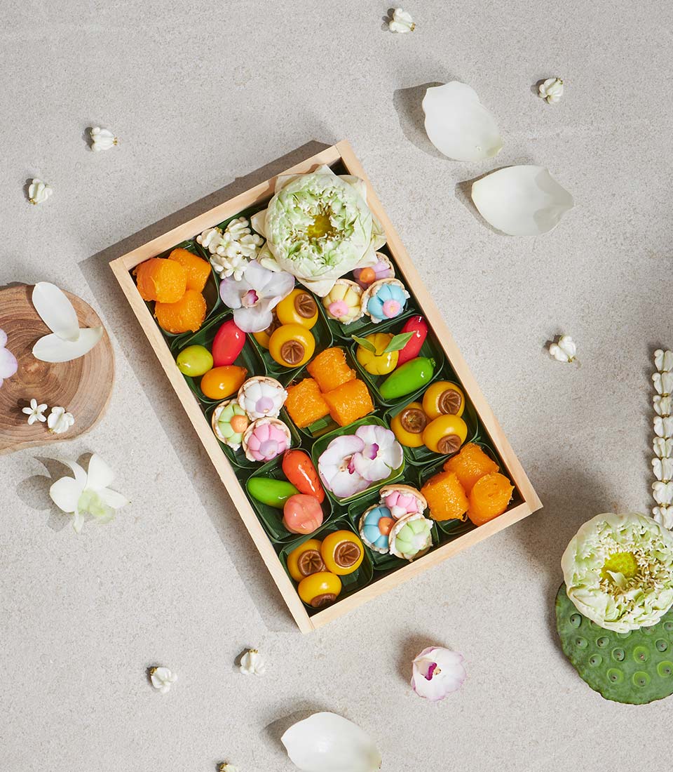 Wooden box of vibrant Thai lotus and jasmine desserts in banana leaf cups with fresh white flowers