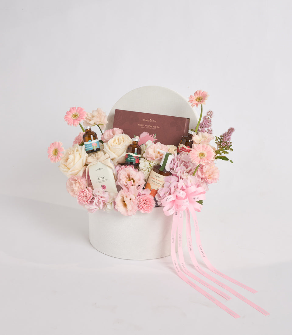 Luxe floral gift box arrangement with pink gerberas, cream roses, carnations, skincare bottles, burgundy book, white cylindrical box, pink ribbon bows on white background.