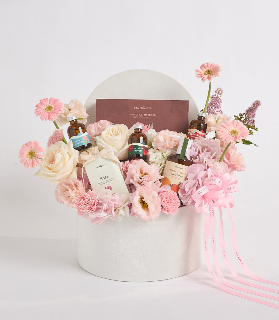Luxurious floral gift set in white cylindrical box with pink gerberas, cream roses, carnations, beauty products, and pink ribbons on white background.