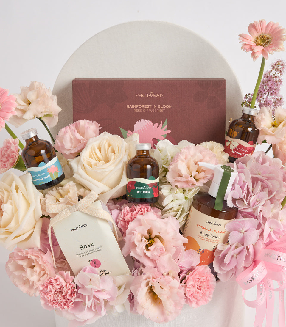 Phutawan luxury beauty gift set surrounded by pink and cream flowers, featuring reed diffuser, body lotion, and skincare products in amber glass bottles.