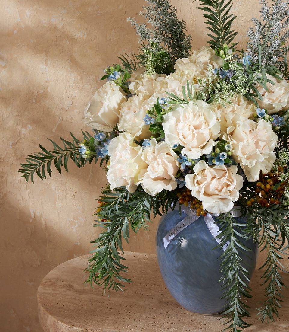 Cream roses and carnations with blue flowers in blue glass vase, premium fresh arrangement