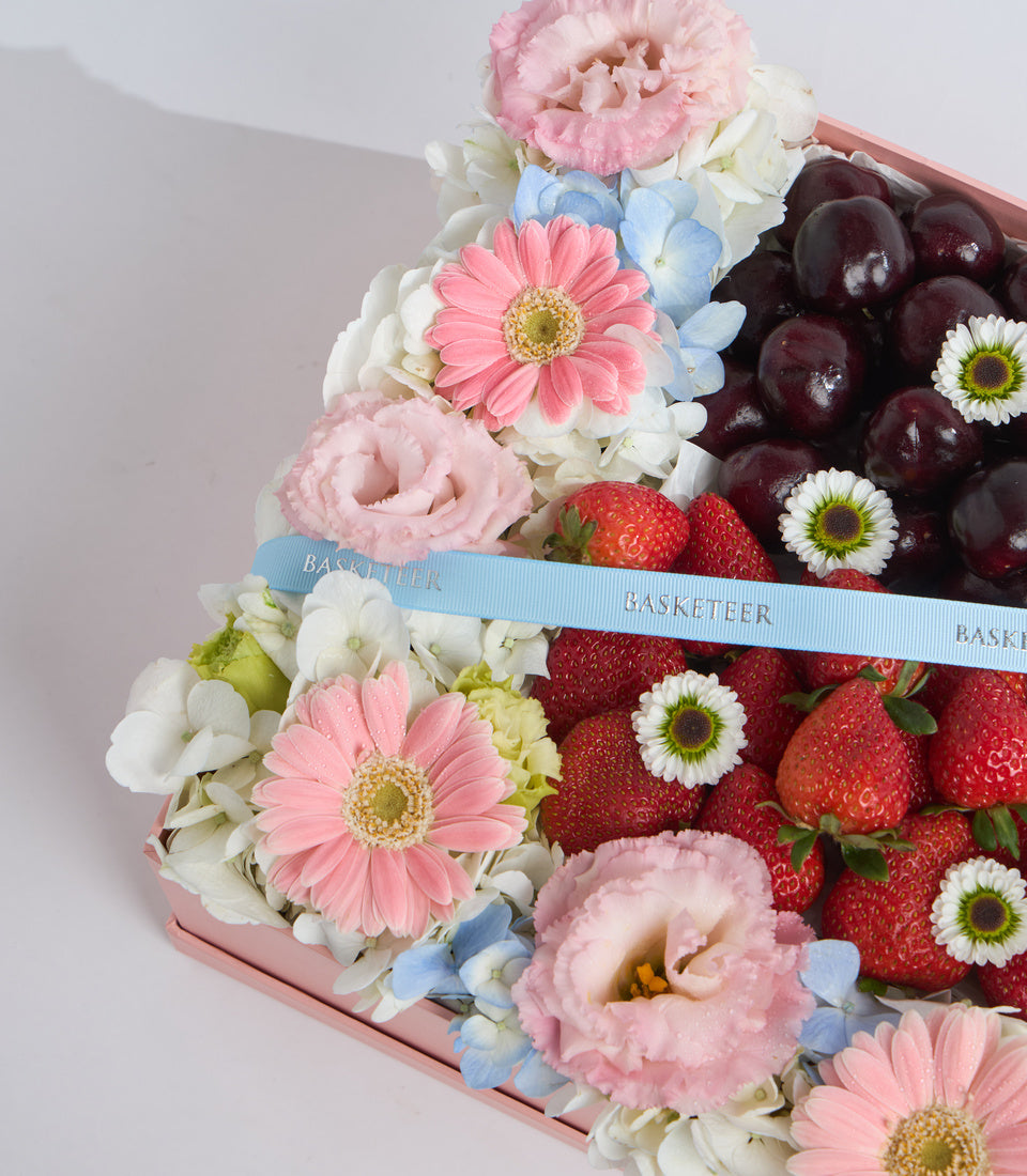 Pink gift box with fresh strawberries, dark grapes, pink peonies, pink gerberas, white flowers, blue hydrangeas, and blue ribbon bow.