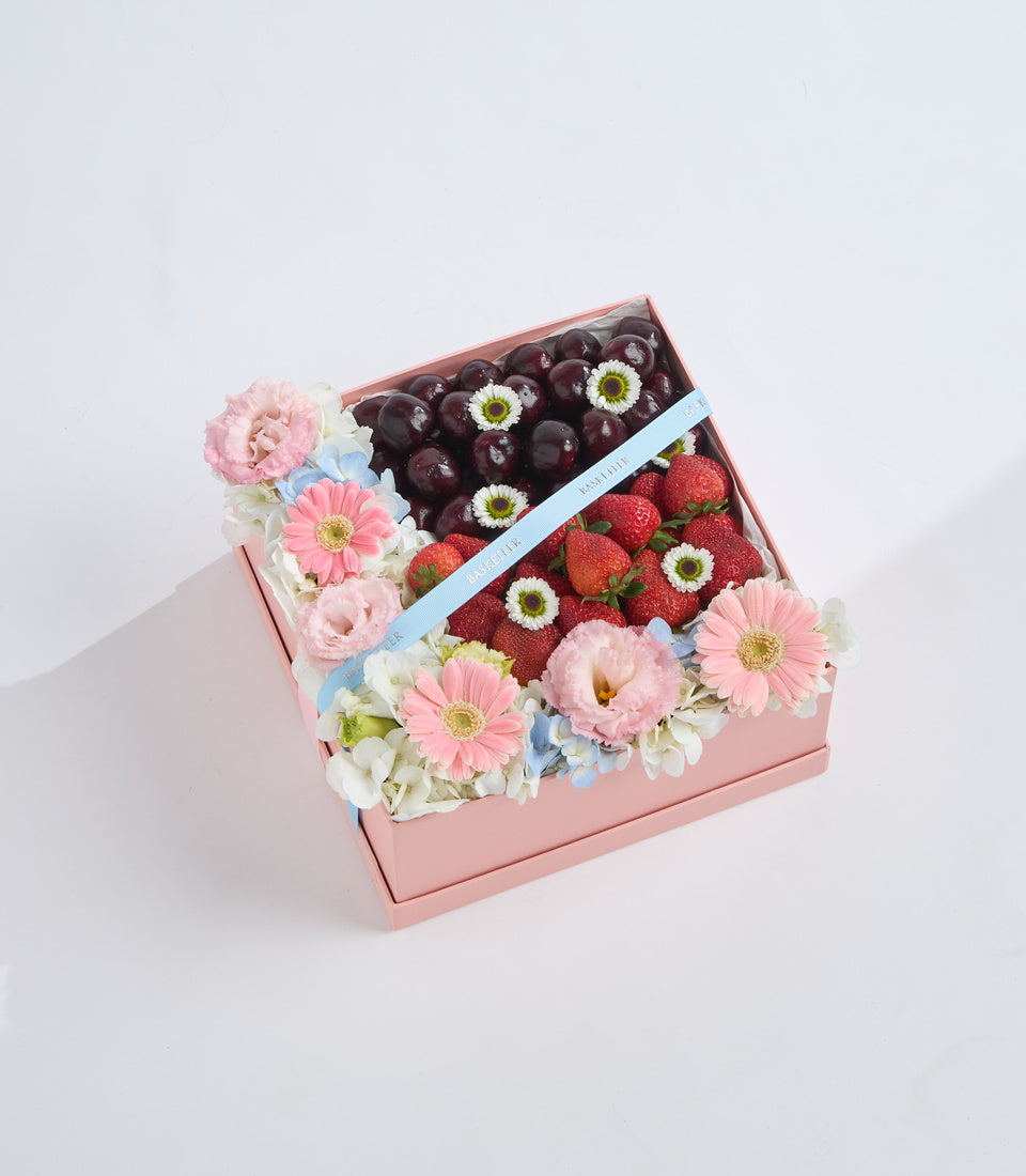 Pink gift box containing dark cherries, fresh strawberries, and pink gerbera daisies with white flowers and blue ribbon accent.