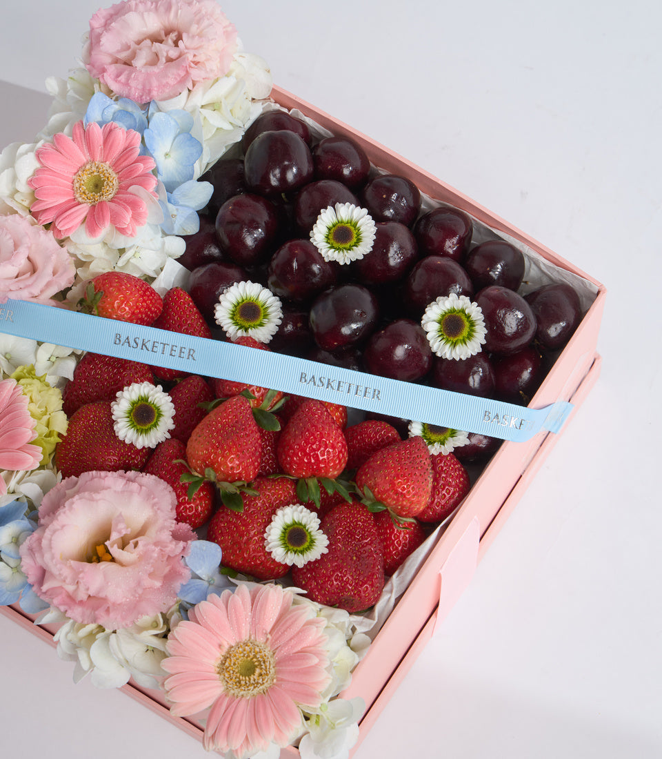 Luxury gift box with fresh strawberries and dark grapes, decorated with pink peonies, gerberas, blue hydrangeas, and white daisies, tied with blue BASKETEER ribbon.