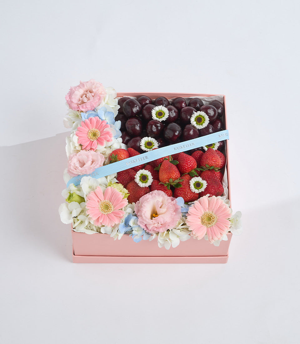 Pink gift box with fresh flowers, strawberries, and dark grapes arranged with white daisies and blue ribbon accent.