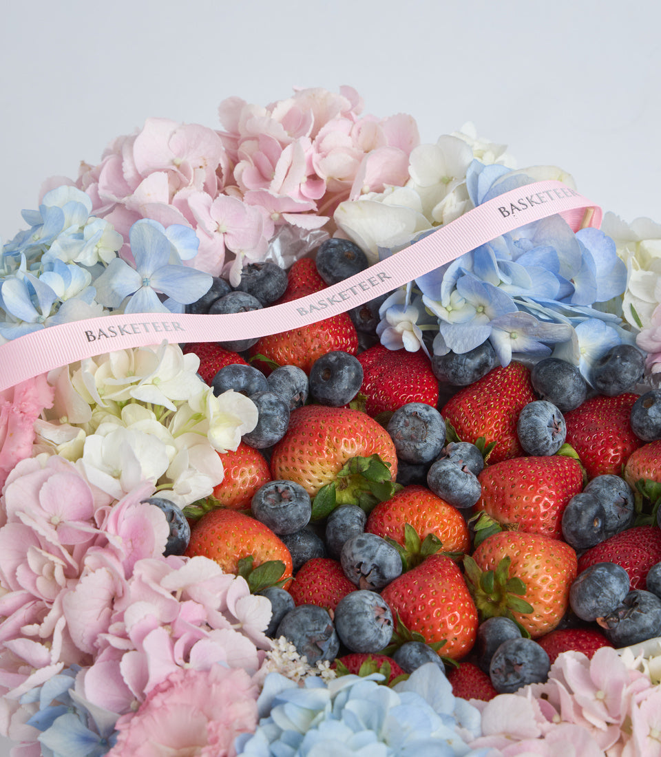Luxury flower and fruit arrangement with pink and blue hydrangeas, fresh strawberries and blueberries, tied with pink "Basketeer" ribbon on white background.