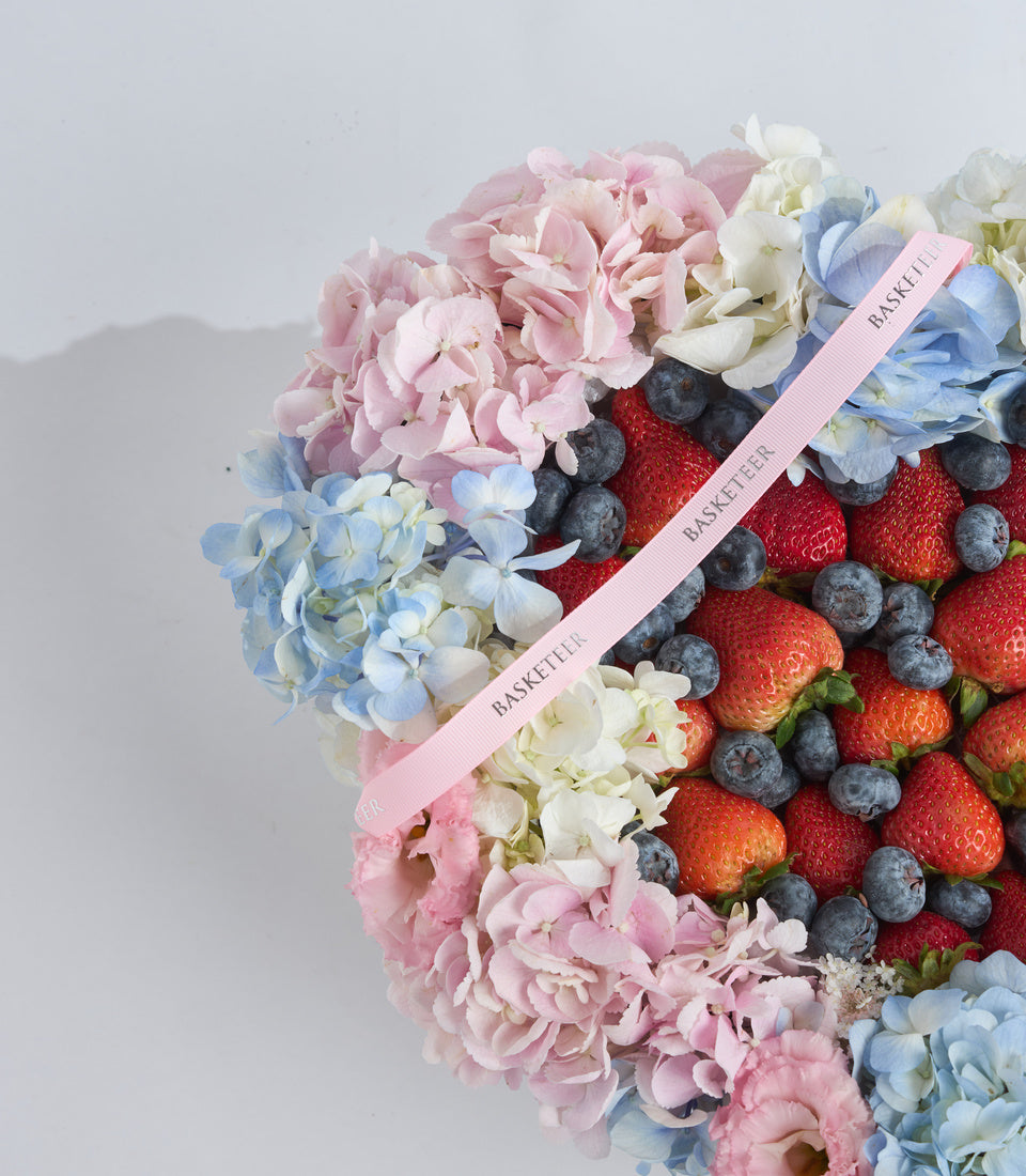 Luxury gift basket with pink ribbon featuring fresh strawberries, blueberries, and pastel pink, blue, and white flowers arranged together.