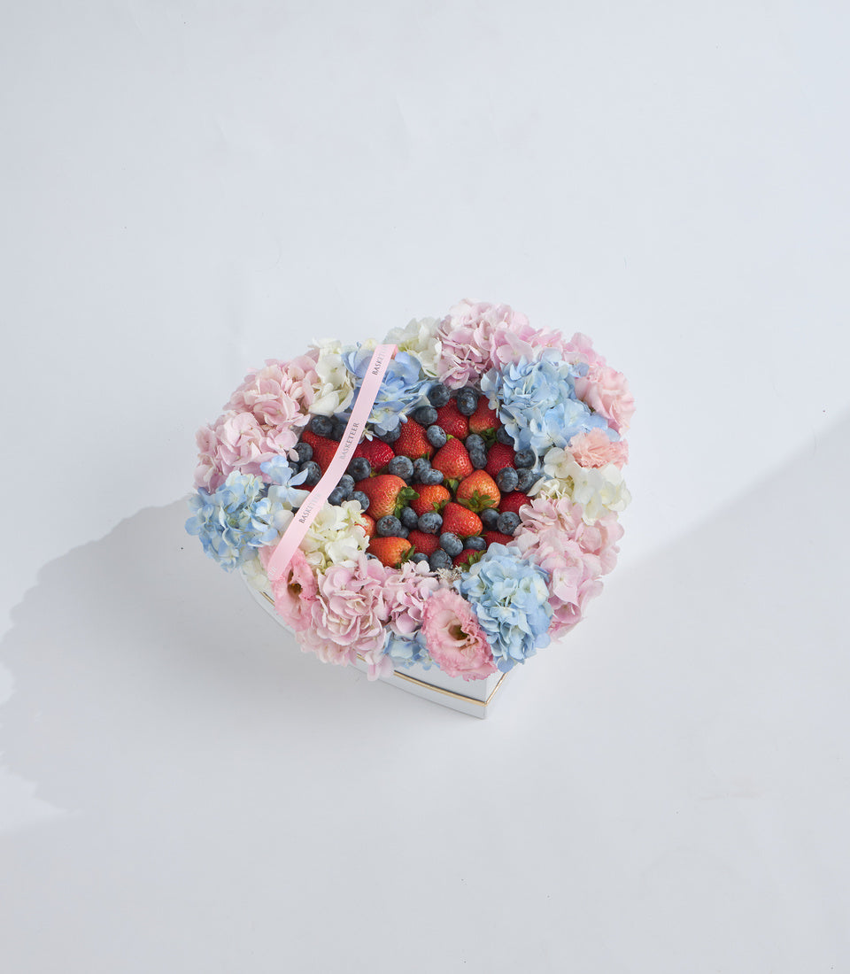 Heart-shaped floral arrangement with pink and blue hydrangeas, white flowers, fresh berries, and pink ribbon on white background.