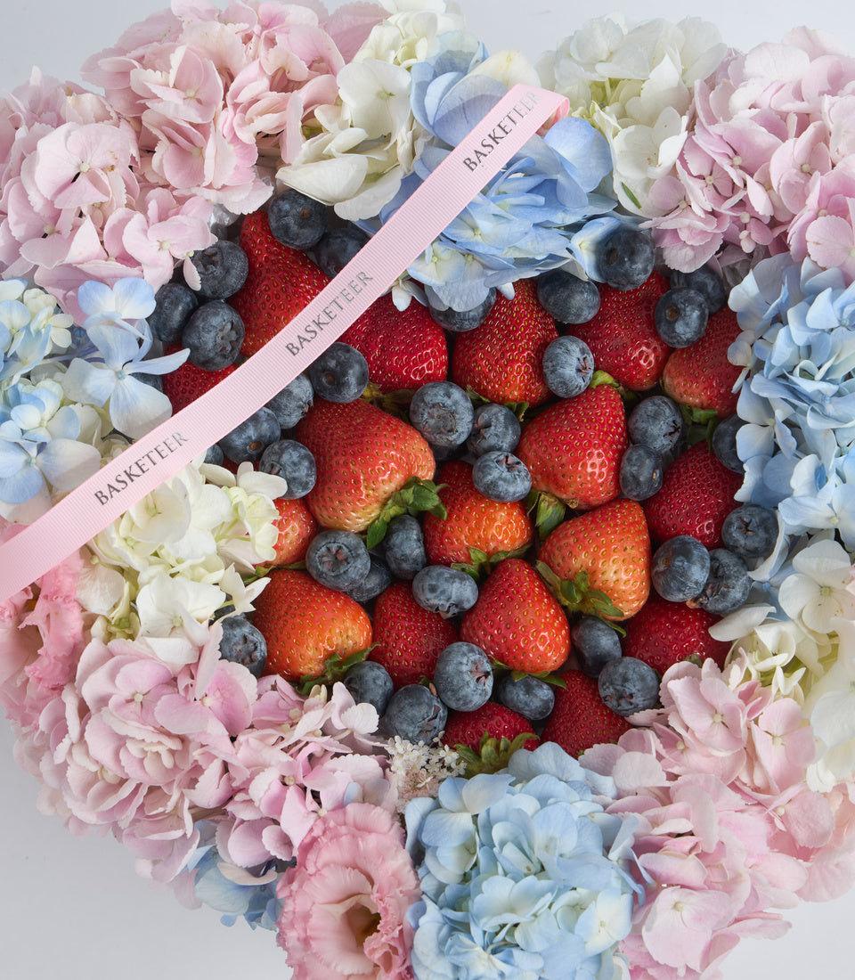 Heart-shaped luxury gift basket with fresh strawberries and blueberries surrounded by pink, white, and blue hydrangeas and carnations.
