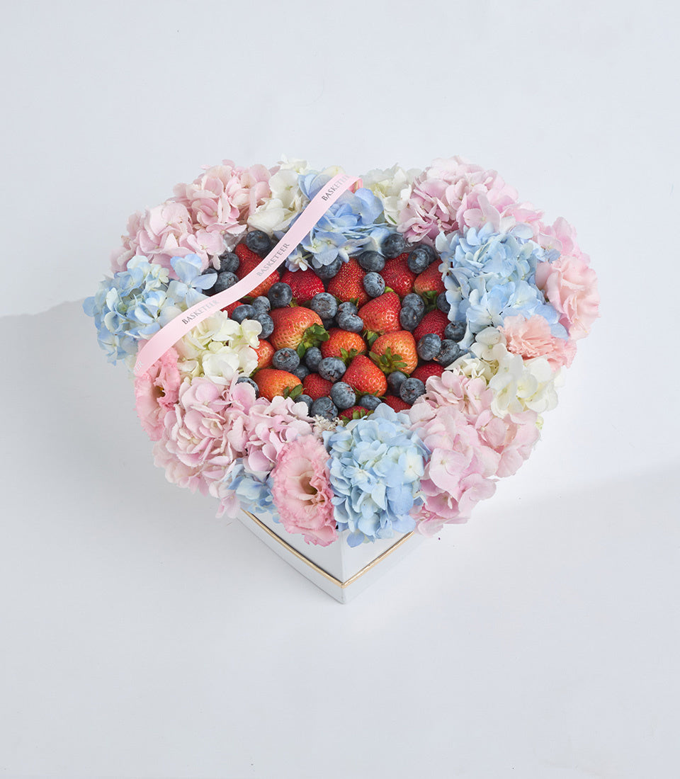 Heart-shaped floral box with pink and blue hydrangeas, carnations, fresh strawberries and blueberries, topped with pink ribbon.