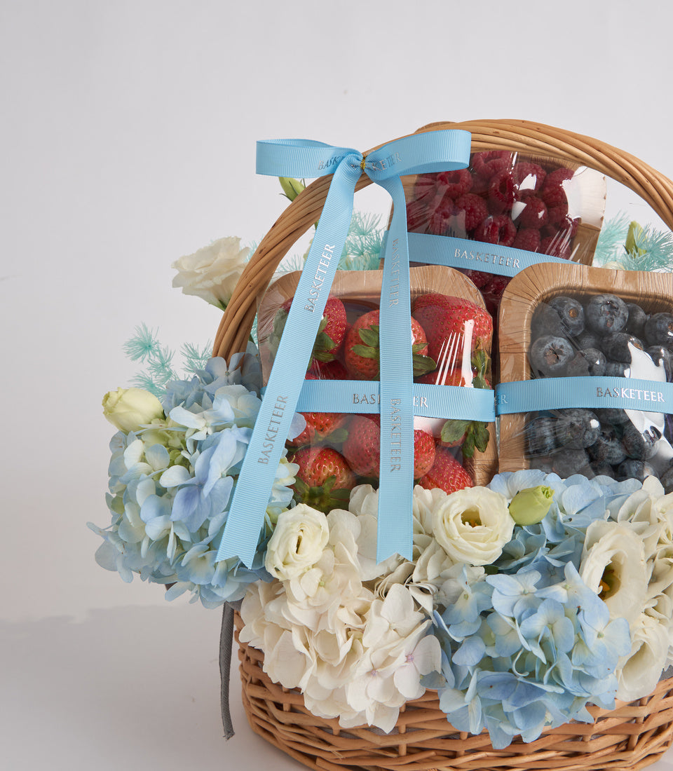 Wicker basket with fresh flowers, strawberries, and blueberries, tied with light blue ribbon bow, featuring white roses and hydrangeas.