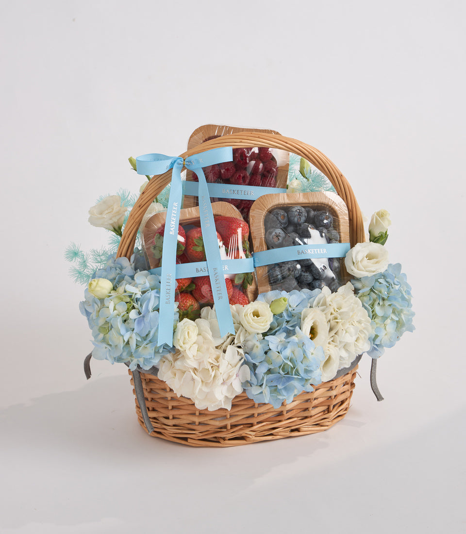 Wicker gift basket with blue and white flowers, fresh strawberries, dark berries, and gourmet items tied with light blue ribbon.