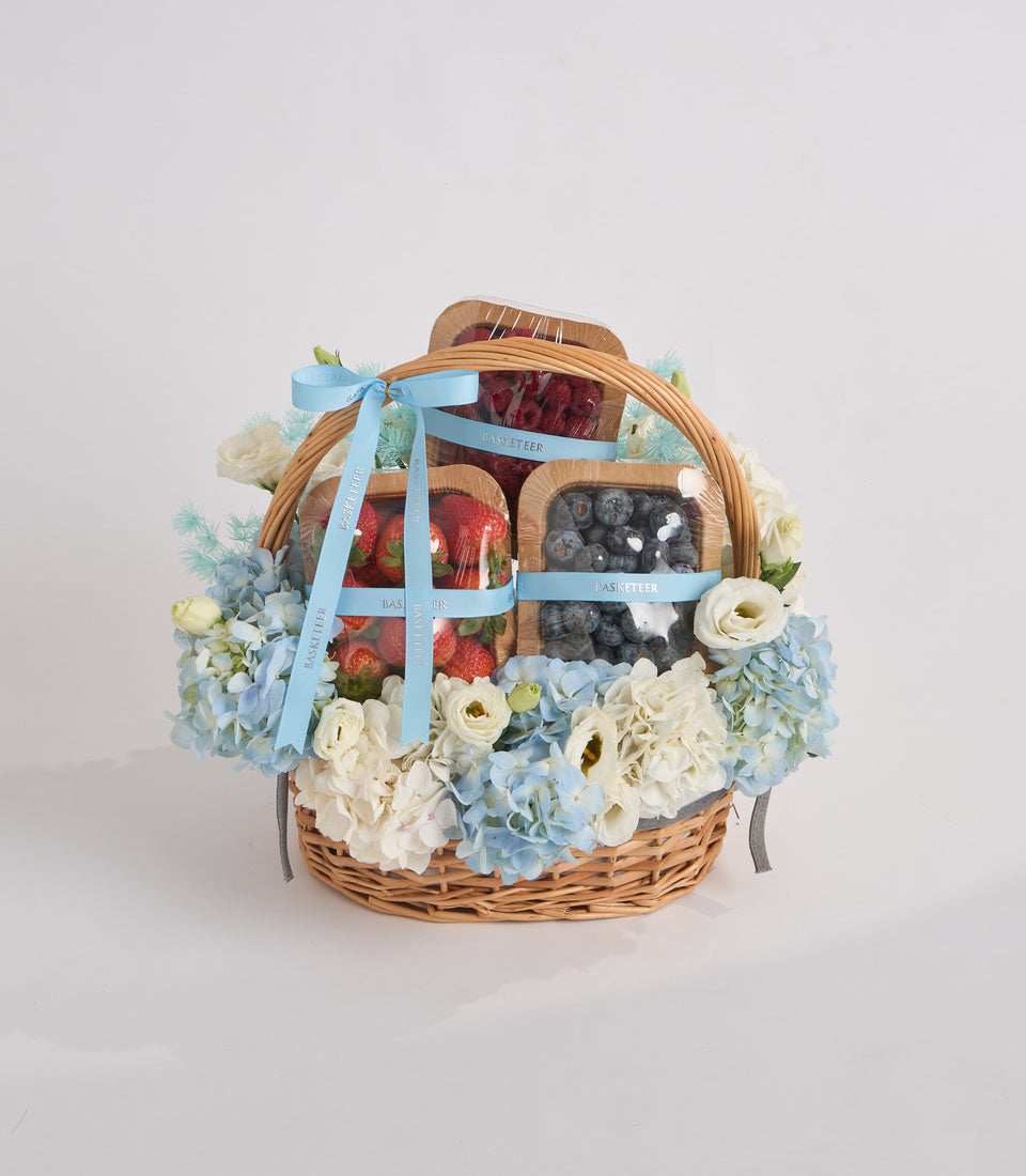 Wicker basket filled with fresh strawberries and blueberries, adorned with white and blue hydrangeas, cream roses, and light blue ribbon bow.