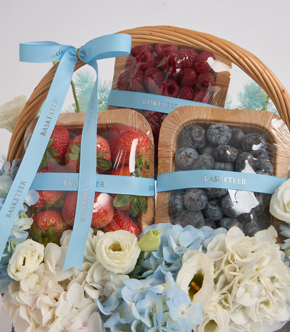 Luxury fruit gift basket with fresh strawberries and blueberries in cardboard containers, decorated with white roses, blue hydrangeas, and light blue ribbon bow.