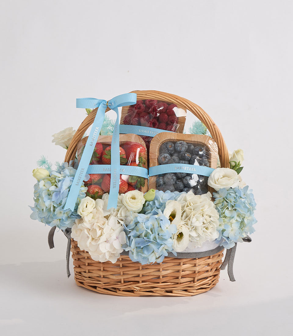Elegant wicker gift basket with fresh berries, blue and white flowers, light blue ribbons, and gray handles on white background.
