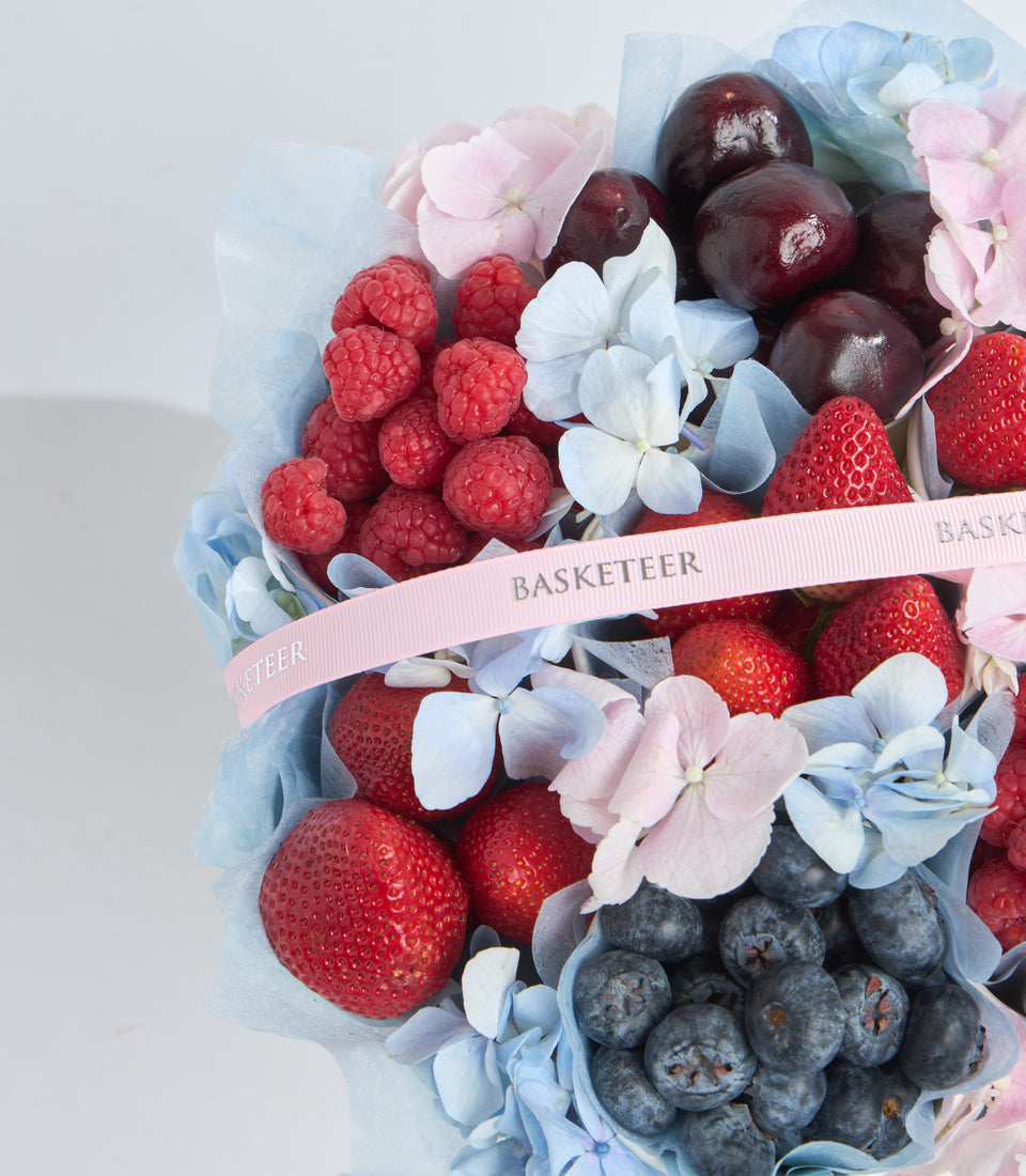 Elegant fruit and flower gift basket with strawberries, raspberries, blueberries, and dark plums, accented with pink and blue hydrangeas, wrapped in tissue paper with pink "Basketeer" ribbon.