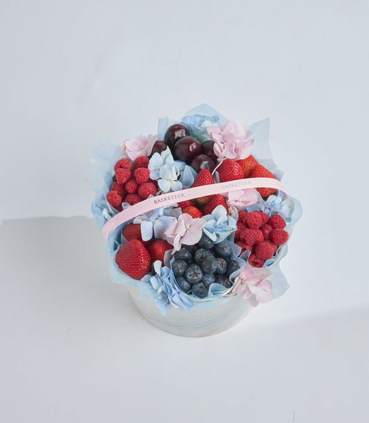 # Alt Text

Elegant fruit and flower arrangement in white basket with fresh berries, pink hydrangeas, blue flowers, and pink ribbon bow on white background.