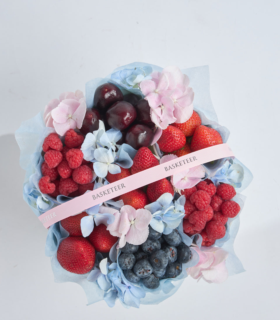 # Alt Text

Elegant fruit and flower gift bouquet featuring strawberries, raspberries, blueberries, plums, pink hydrangeas, and blue flowers wrapped in translucent paper with pink "Basketeer" ribbon.
