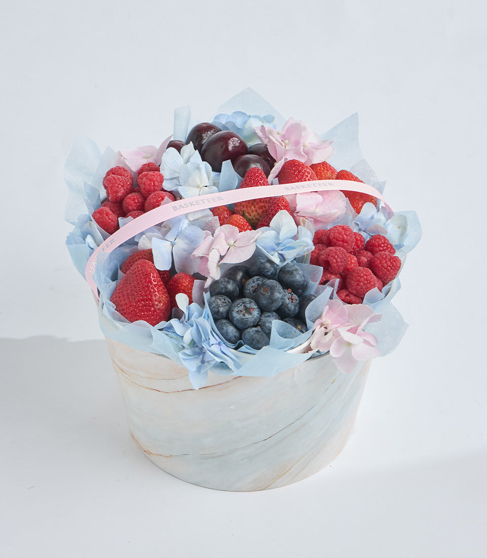 # Alt Text

Luxury fruit bouquet with strawberries, raspberries, blueberries, and dark cherries, arranged with blue and pink flowers in a white textured box with pink ribbon.