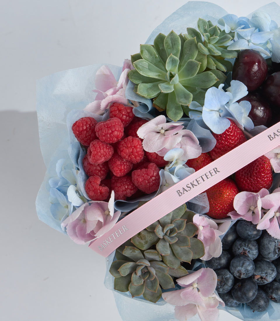 Luxurious gift basket with fresh raspberries, strawberries, blueberries, succulent plants, blue and pink hydrangeas, wrapped in light blue paper with pink ribbon.