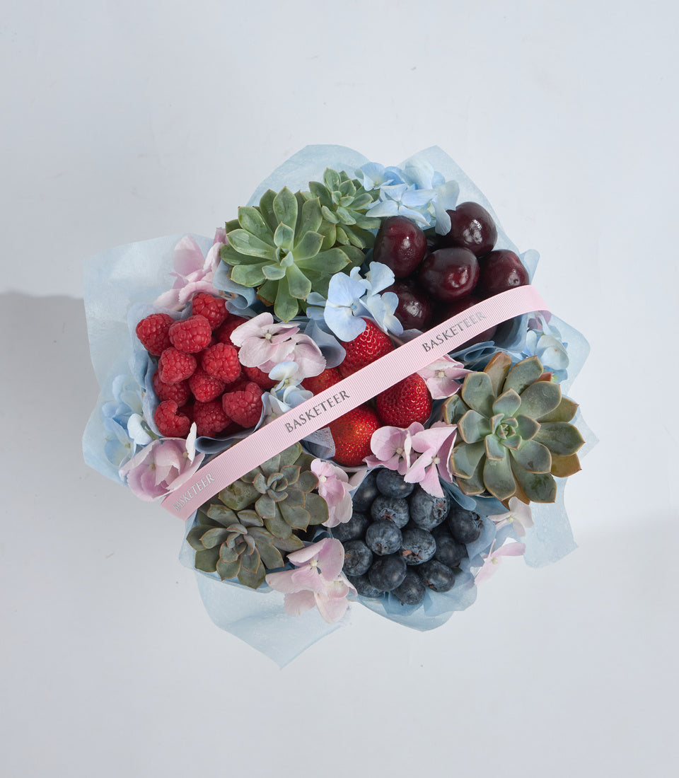 Luxury fruit and succulent arrangement with raspberries, blueberries, cherries, strawberries, blue flowers, green succulents, wrapped in blue cellophane with pink ribbon.